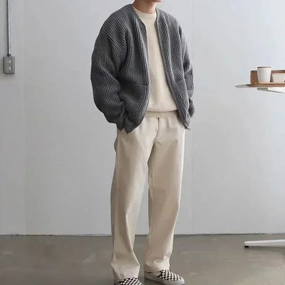 Person wearing a gray cardigan, beige sweater, and beige pants in a minimalistic room.