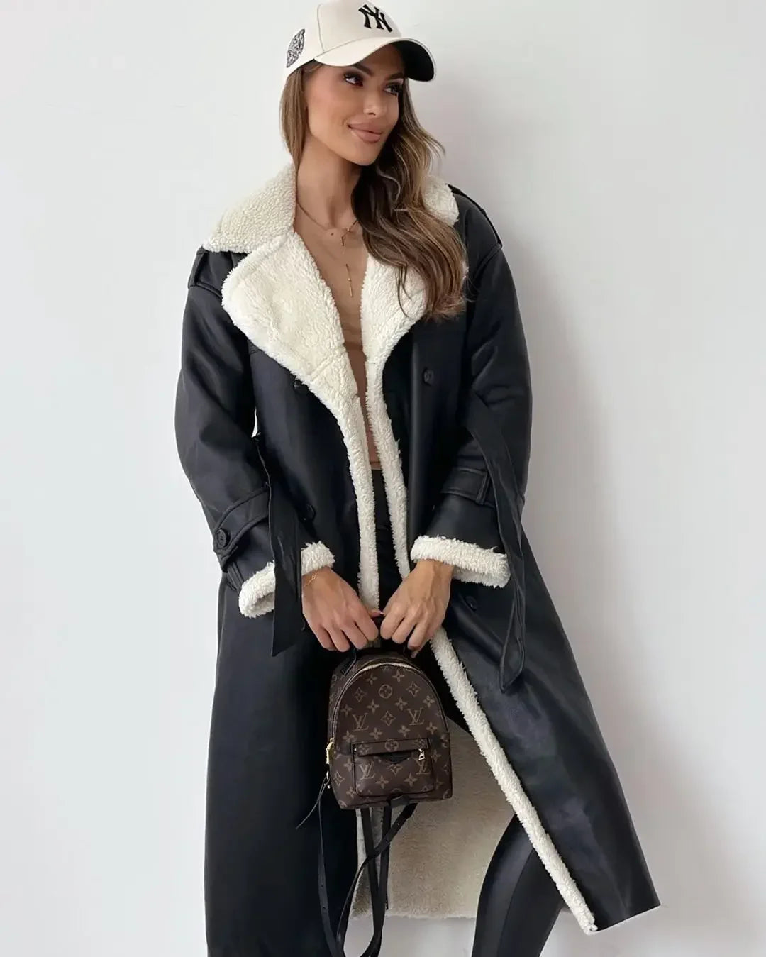 Woman wearing a black coat with white fur lining, holding a brown handbag against a white background