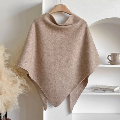 Beige poncho on a wooden hanger against a white wall with a shelf in the background.