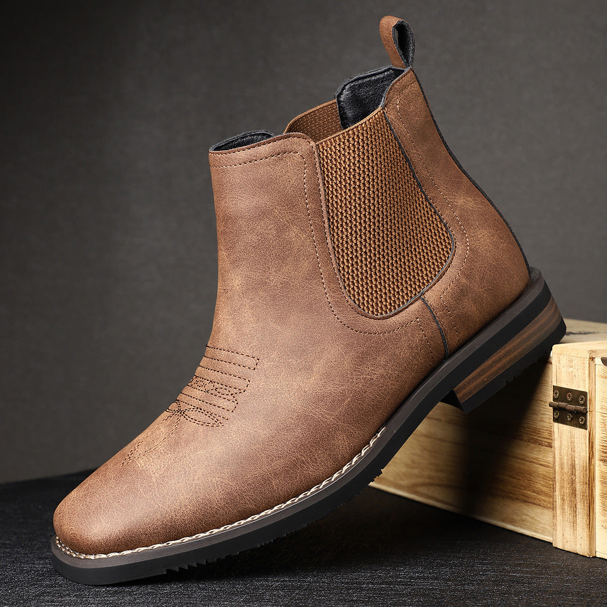 Brown leather Chelsea boot on a dark surface with a wooden box in the background