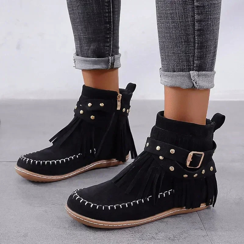 Black fringe boots with gold studs and buckles worn with rolled-up jeans on a light gray background
