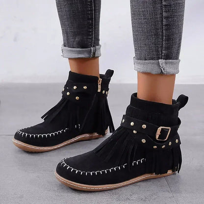 Black fringe boots with gold studs and buckles worn with rolled-up jeans on a light gray background