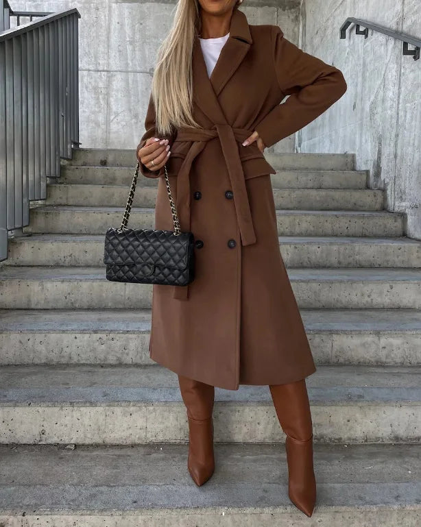 Woman in a brown coat and boots holding a black quilted handbag on concrete steps.