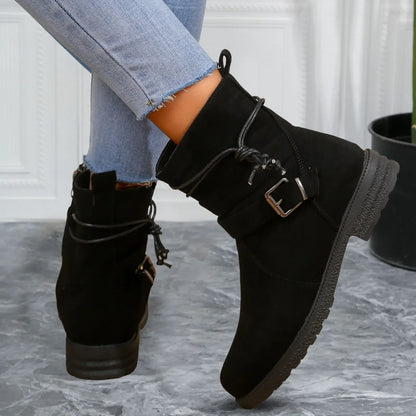Black suede ankle boots with a buckle detail worn with light blue jeans on a gray surface.