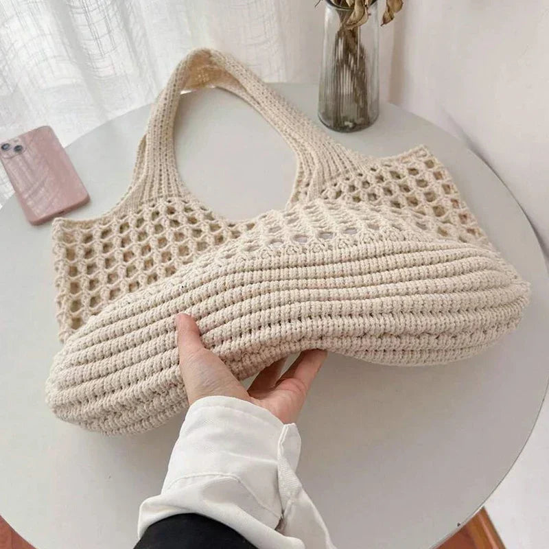 Beige woven handbag held by a person on a white surface with a phone and vase in the background.