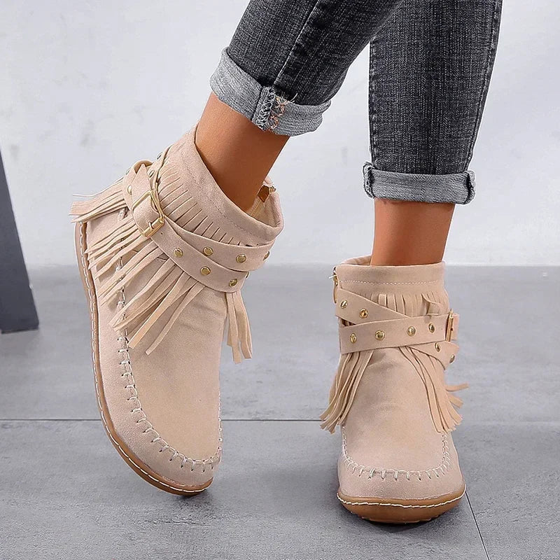 Beige fringed boots worn with rolled-up jeans on a gray floor.
