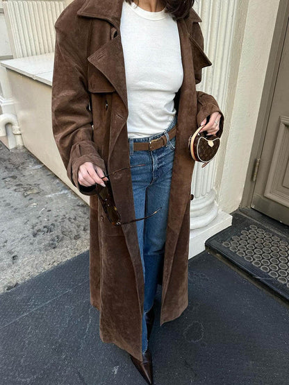 Person wearing a brown coat, white shirt, and blue jeans standing on a doorstep.