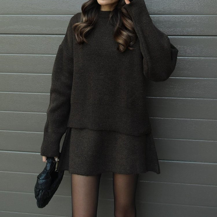Person wearing a dark gray sweater and skirt set against a gray wall.