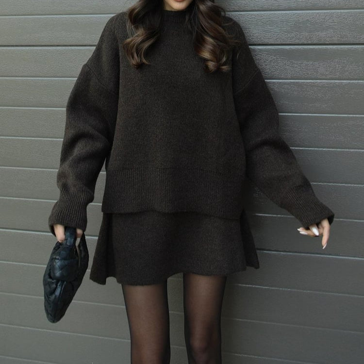 Person wearing a dark gray sweater and skirt set against a gray wall.