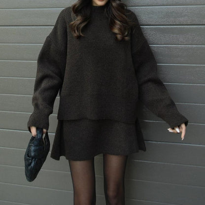 Person wearing a dark gray sweater and skirt set against a gray wall.