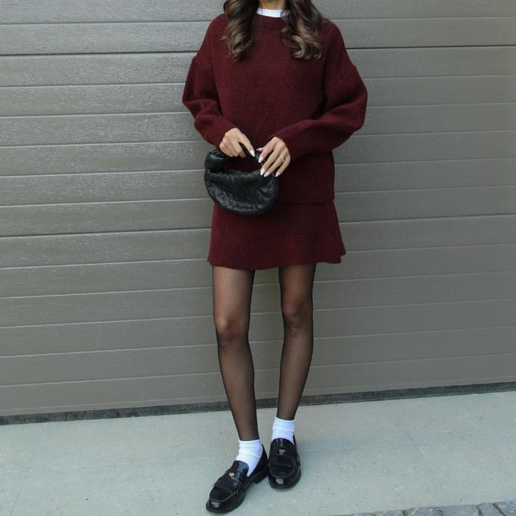 Person wearing a burgundy sweater dress and black shoes against a gray wall.