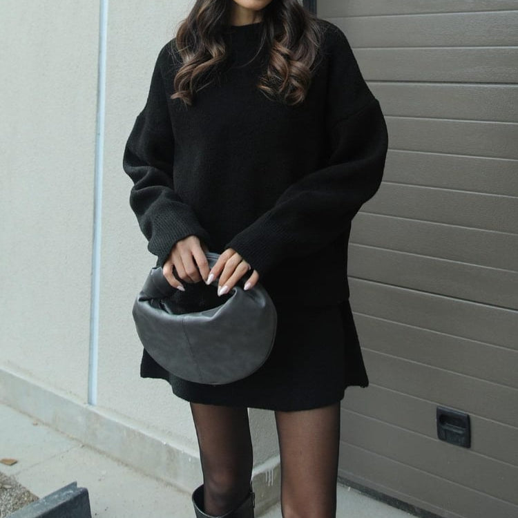 Person wearing a black oversized sweater and holding a gray helmet against a plain wall.