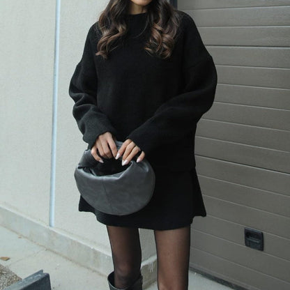 Person wearing a black oversized sweater and holding a gray helmet against a plain wall.