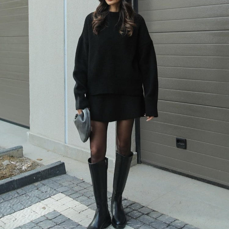 Person wearing a black outfit with knee-high boots standing against a gray wall.