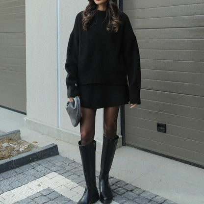 Person wearing a black outfit with knee-high boots standing against a gray wall.