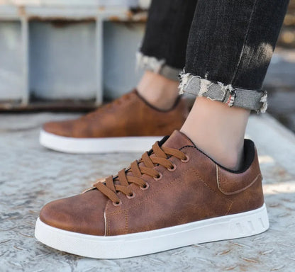 Brown leather sneakers with white soles worn with rolled-up jeans on a concrete surface.