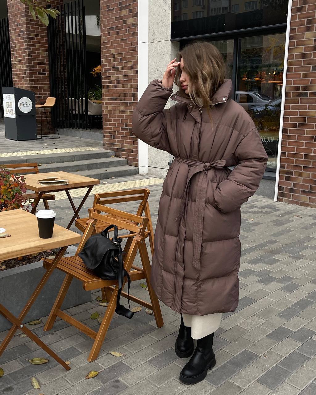 Person wearing a long brown puffer coat standing outdoors near a cafe.