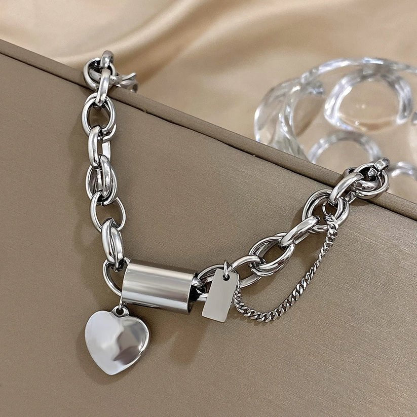 Silver chain bracelet with heart charm and cylindrical tag on a beige surface