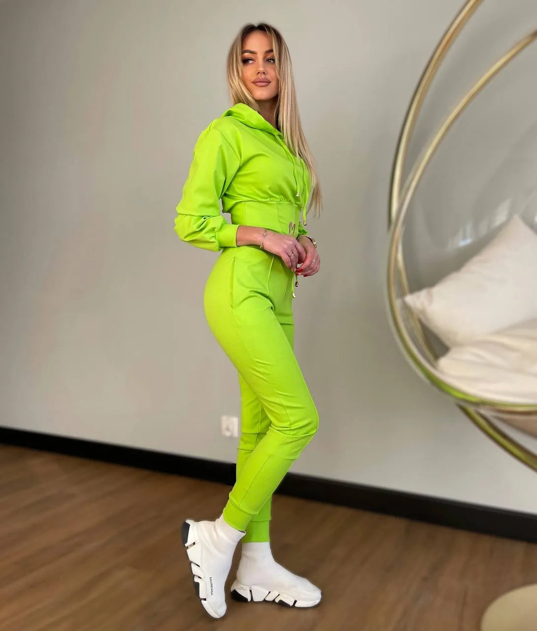 Woman wearing a bright green tracksuit standing in a room with a neutral wall and wooden floor.