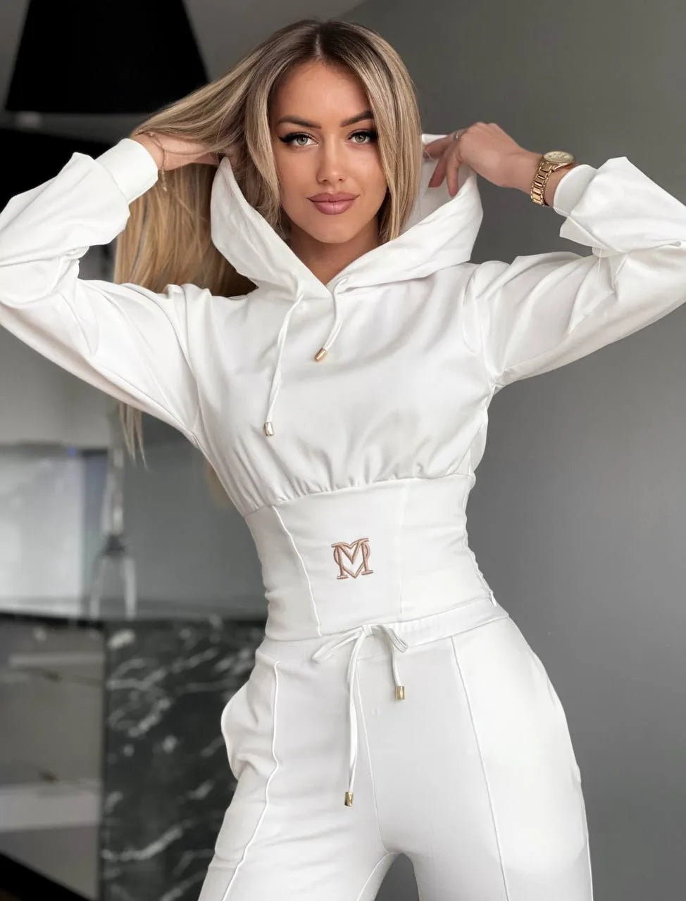 Woman wearing a white hoodie and pants with a logo, standing indoors.