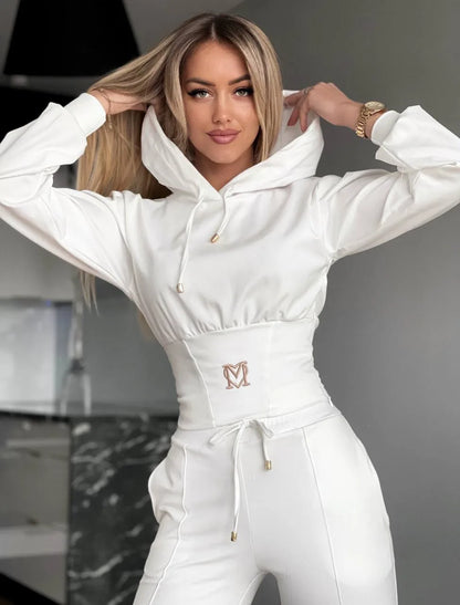 Woman wearing a white hoodie and pants with a logo, standing indoors.