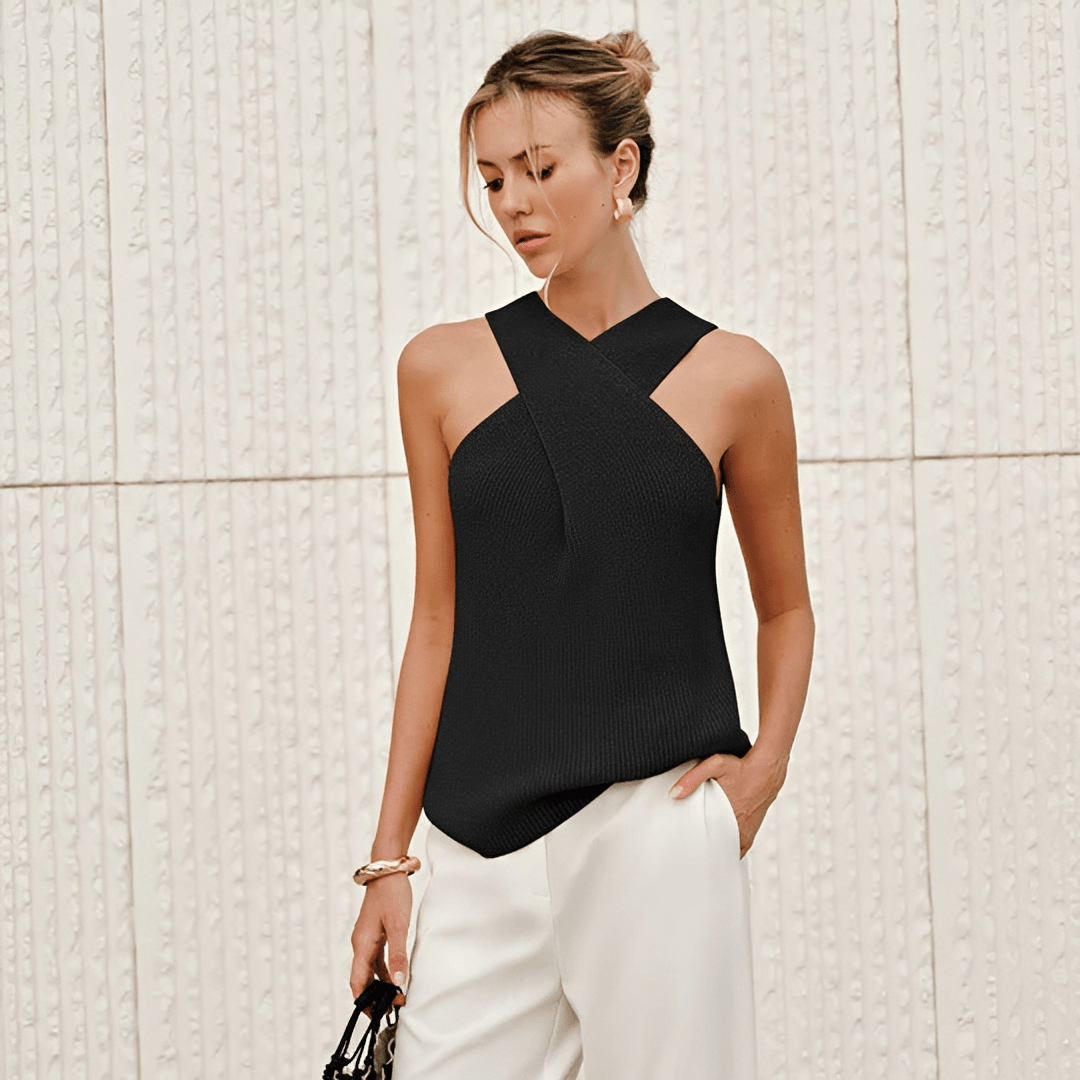 Woman wearing a black sleeveless top and white pants against a light textured wall.