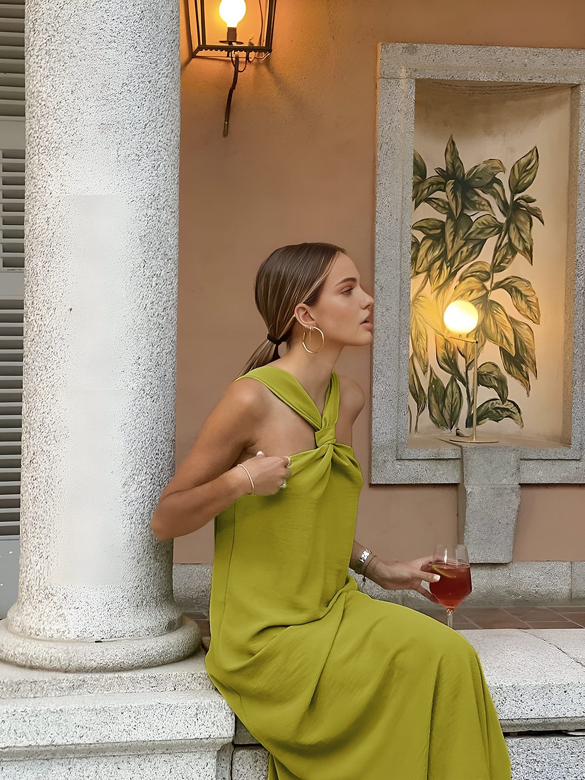 Woman in a green dress holding a glass of red wine, standing against a decorative wall with a plant painting.