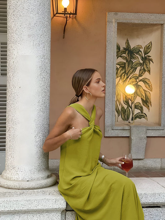 Woman in a green dress holding a glass of red wine, standing against a decorative wall with a plant painting.