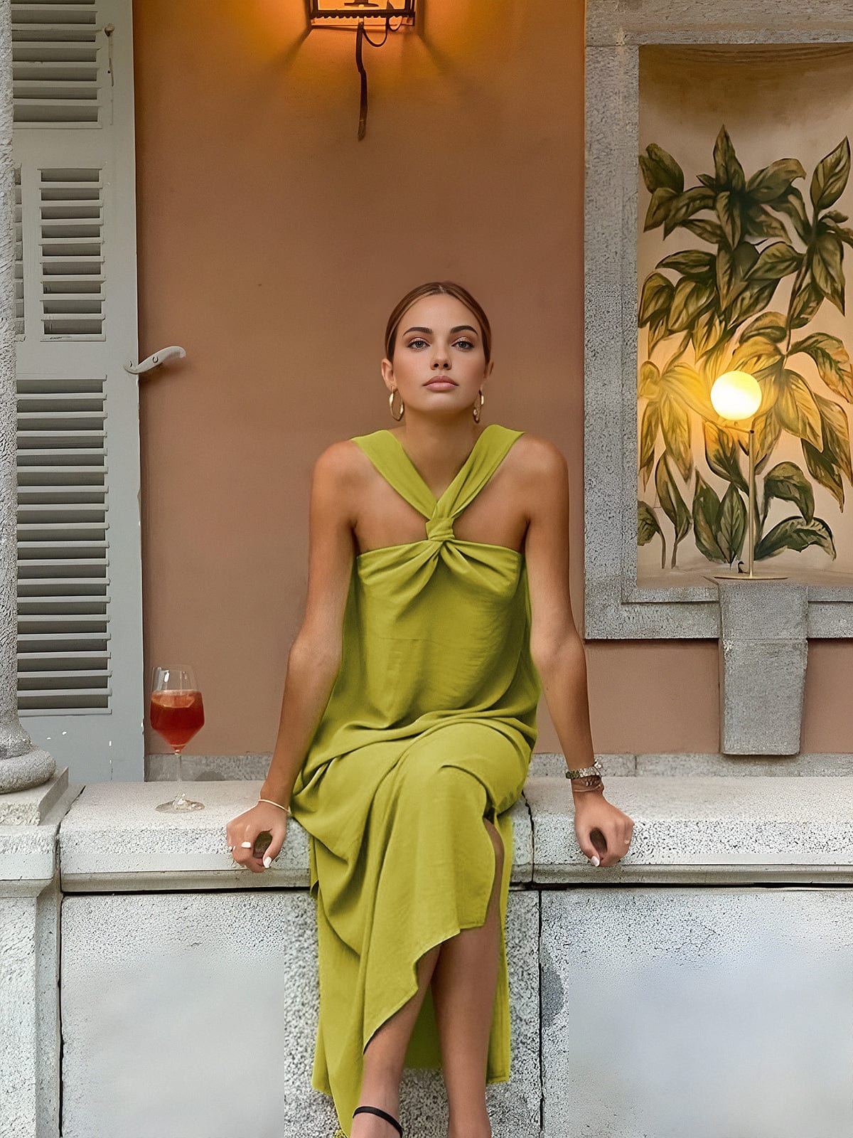 Woman in a green dress sitting on a ledge with a drink, surrounded by warm lighting and decor.