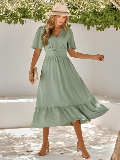 Woman wearing a green dress and straw hat against a white wall with greenery.