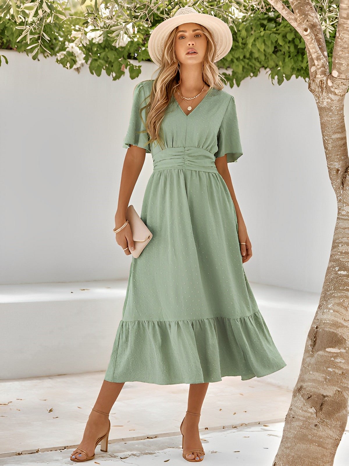 Woman wearing a green dress and hat standing under a tree with a white wall background