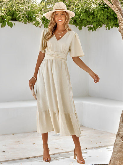 Woman in a beige dress and hat standing in a sunlit outdoor setting with greenery.