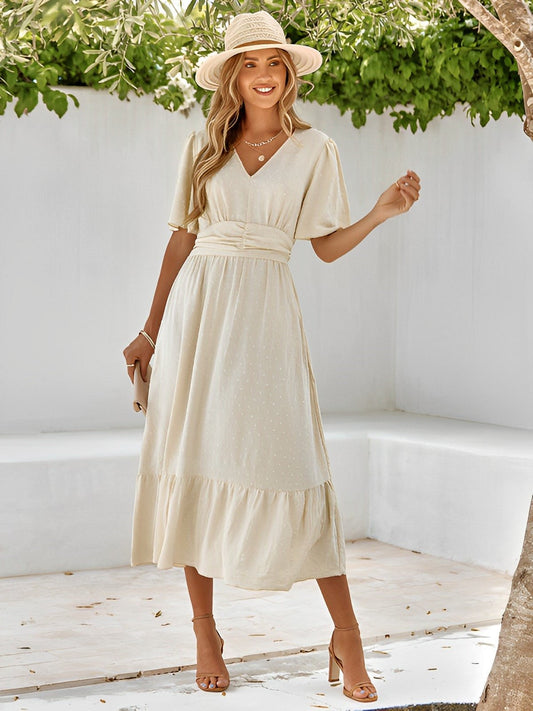 Woman in a beige dress and hat standing in a sunlit outdoor setting with greenery.