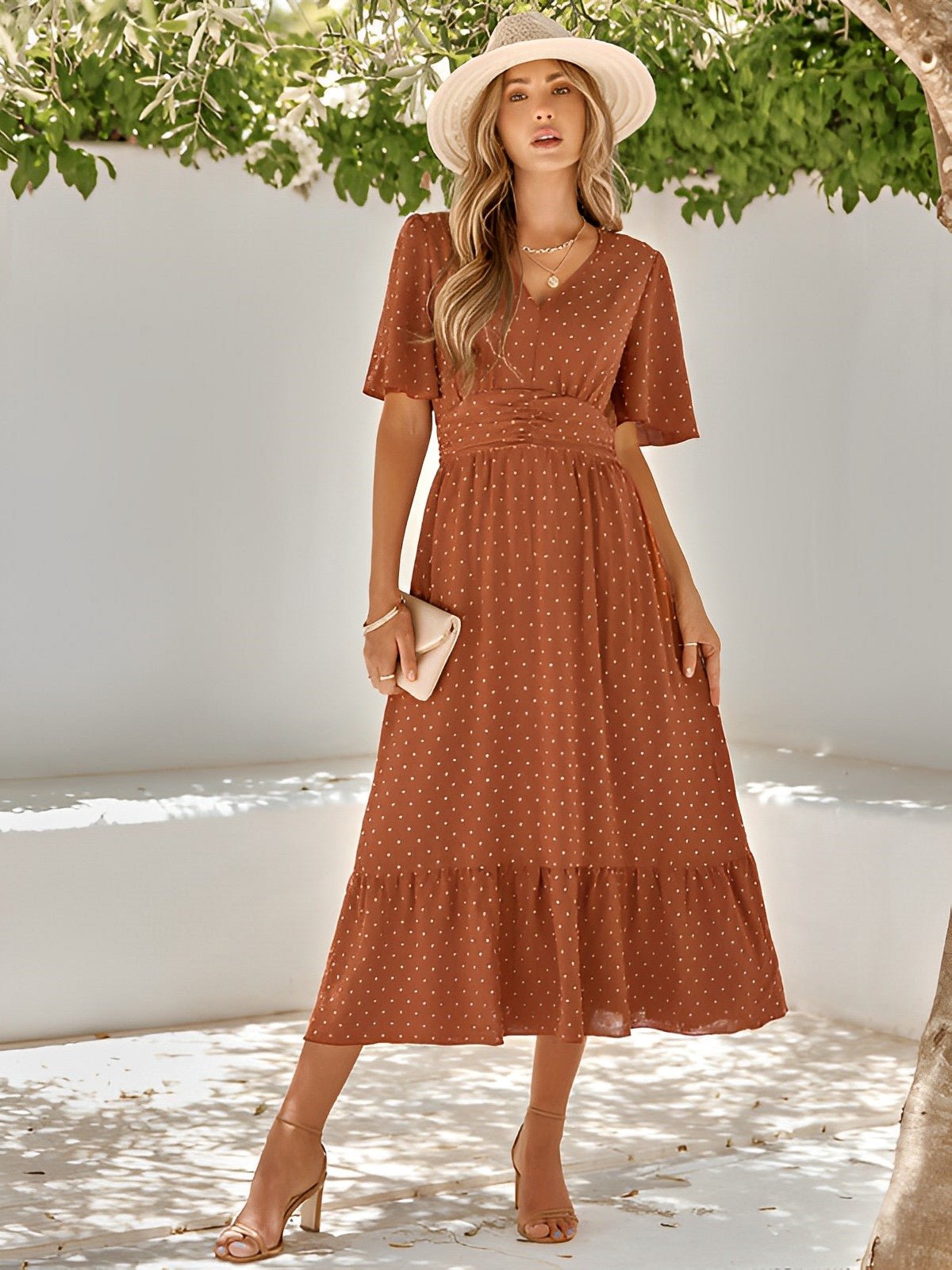Woman wearing a rust-colored polka dot dress and hat, standing under greenery.
