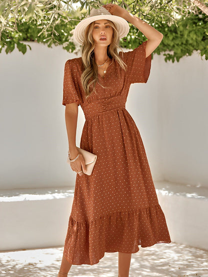 Woman wearing a rust-colored polka dot dress with a white hat, standing against a white wall with greenery.