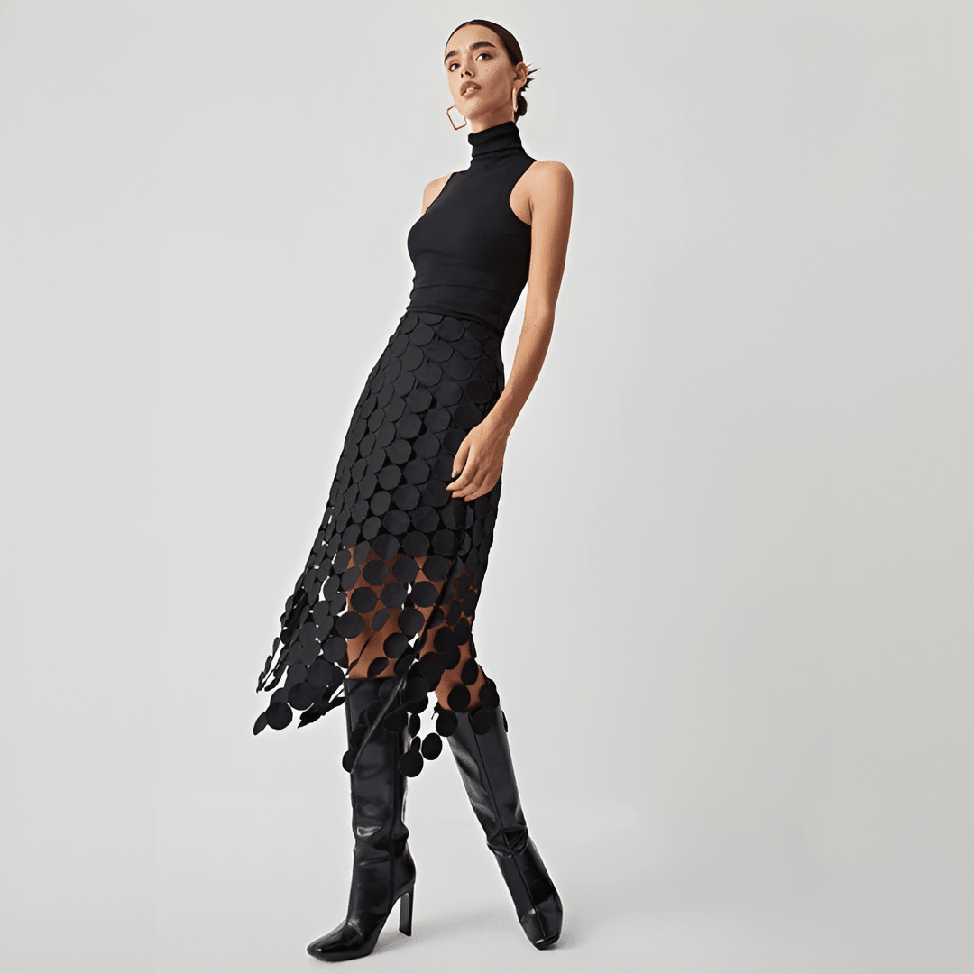Woman wearing a black sleeveless top and black patterned skirt with black knee-high boots on a light gray background