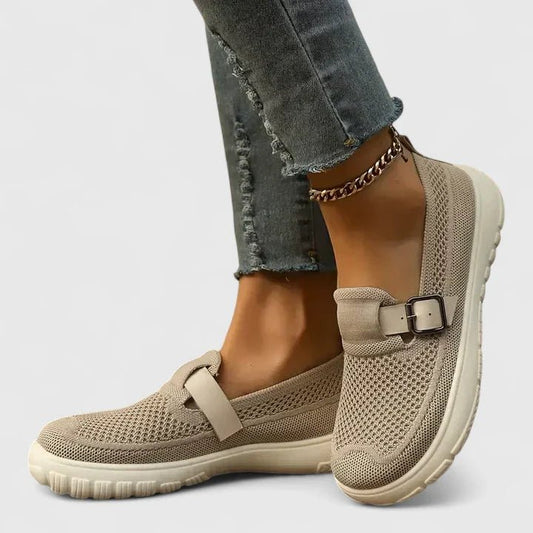 Beige slip-on shoes with a buckle detail worn with ripped jeans on a light gray background