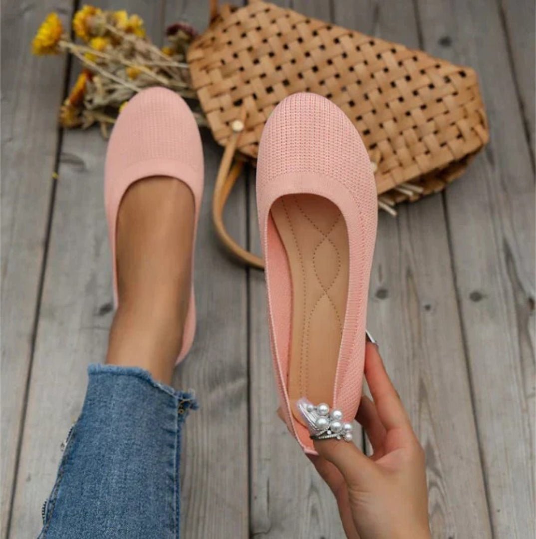 Pink flats held by a hand on a wooden floor with a woven basket and flowers in the background