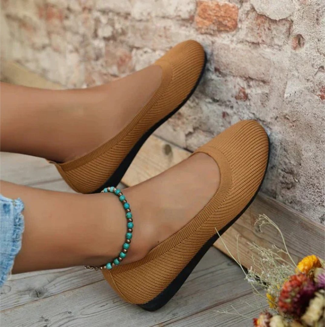 Brown flat shoes worn with denim shorts against a rustic brick wall.