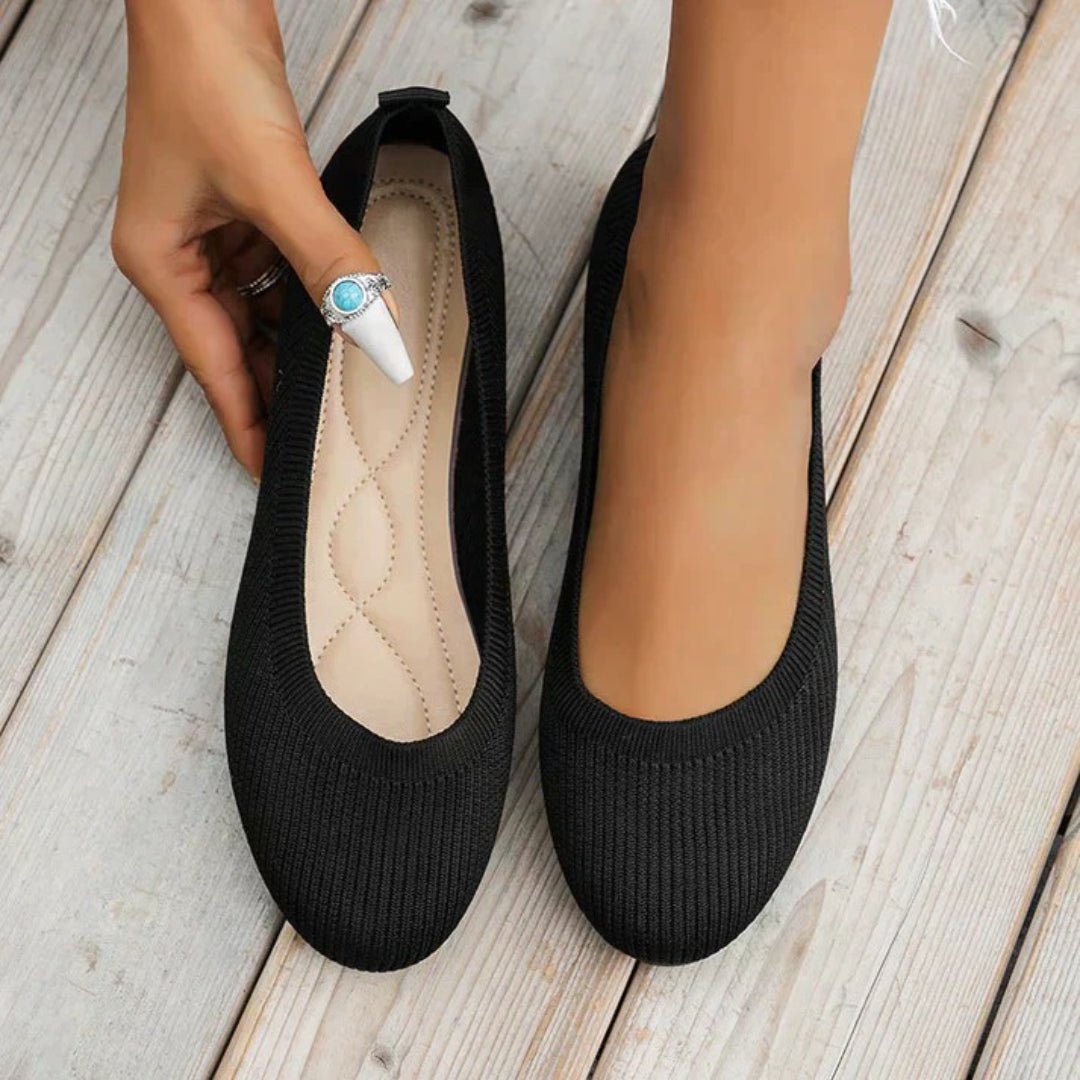 Black flats on a wooden floor with a hand holding one shoe.