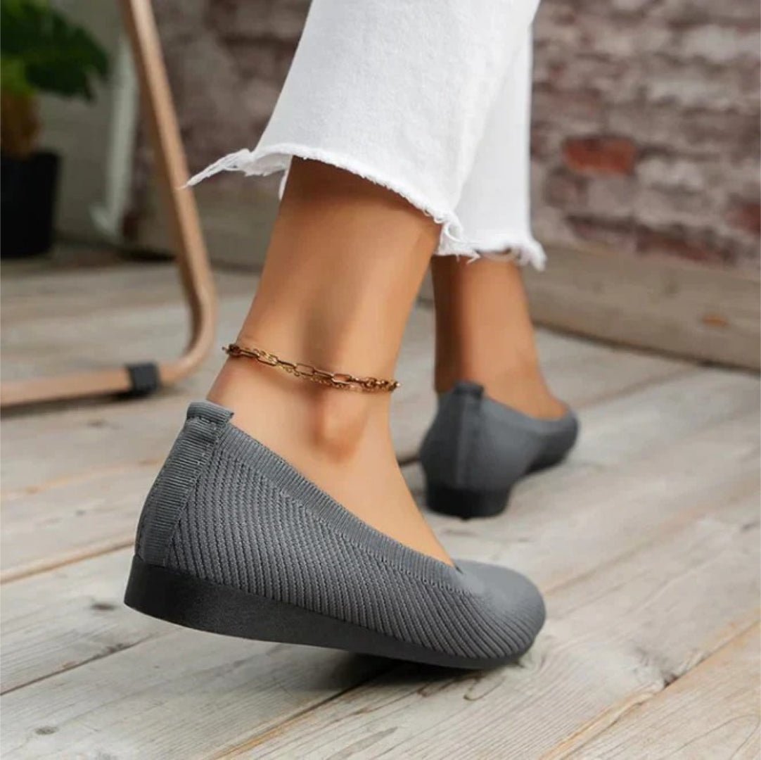 Gray flat shoes worn with white pants on a wooden floor.