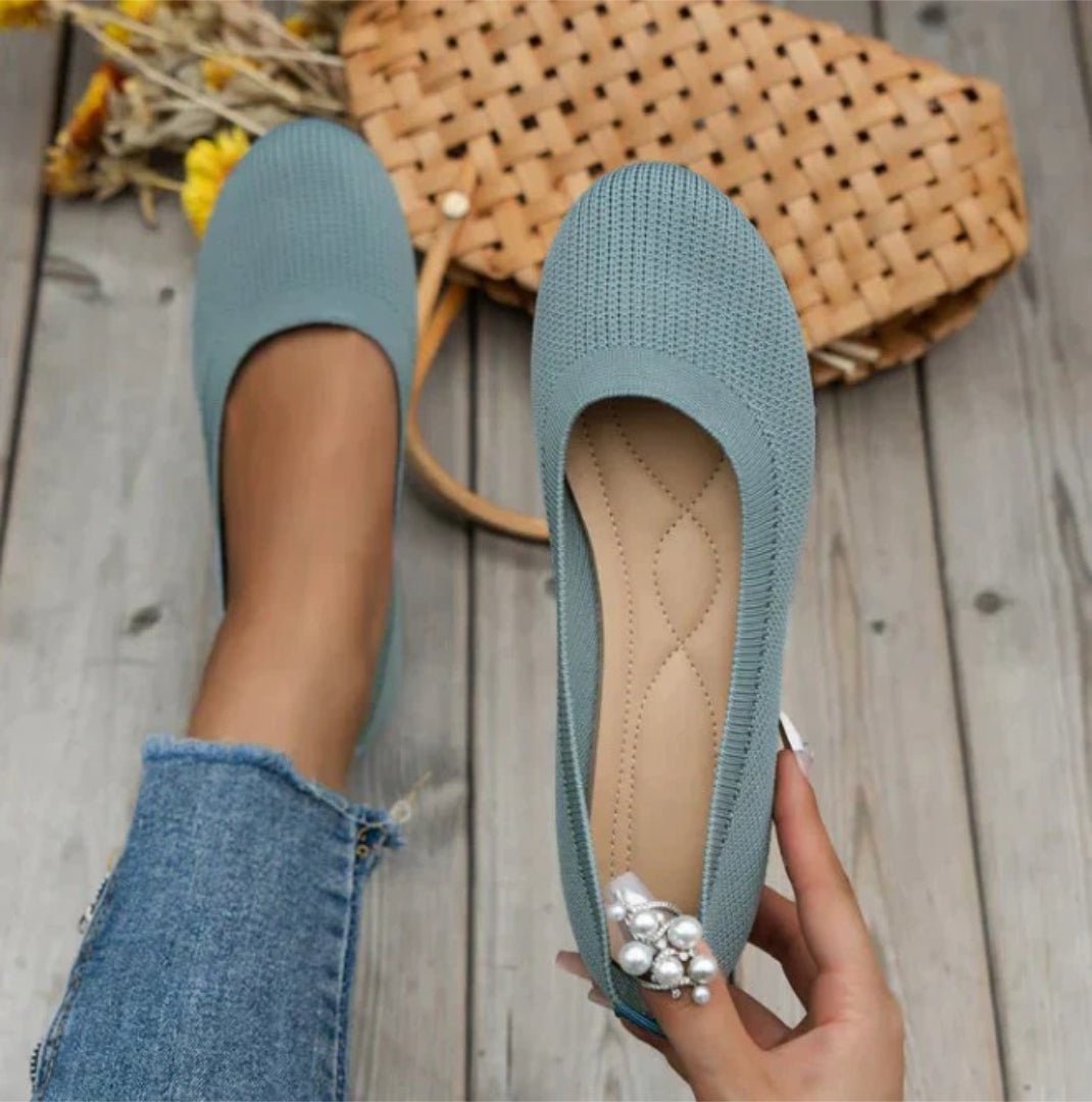 Blue knitted flats with pearl embellishments held by a hand on a wooden floor.