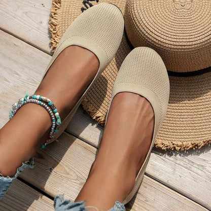 Beige flats worn on a wooden floor with a woven mat and hat in the background.