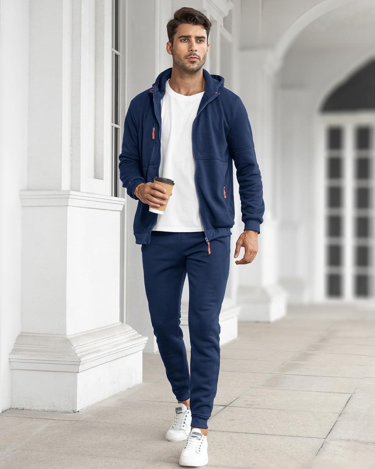 Man wearing a navy tracksuit with a white shirt, holding a coffee cup, standing in a white architectural setting.