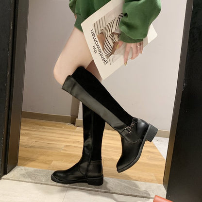 Person wearing black knee-high boots with a magazine in hand, standing on a wooden floor.