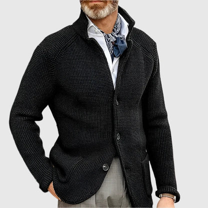 Man wearing a dark textured cardigan over a light shirt with a patterned scarf on a light gray background