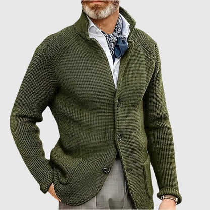 Man wearing a green knitted cardigan over a white shirt with a patterned scarf on a light gray background