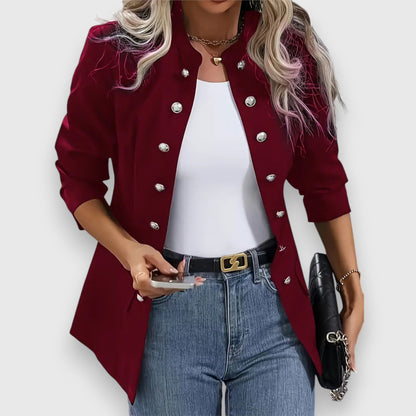 Woman wearing a red blazer with silver buttons, white top, blue jeans, and holding a phone and black clutch.