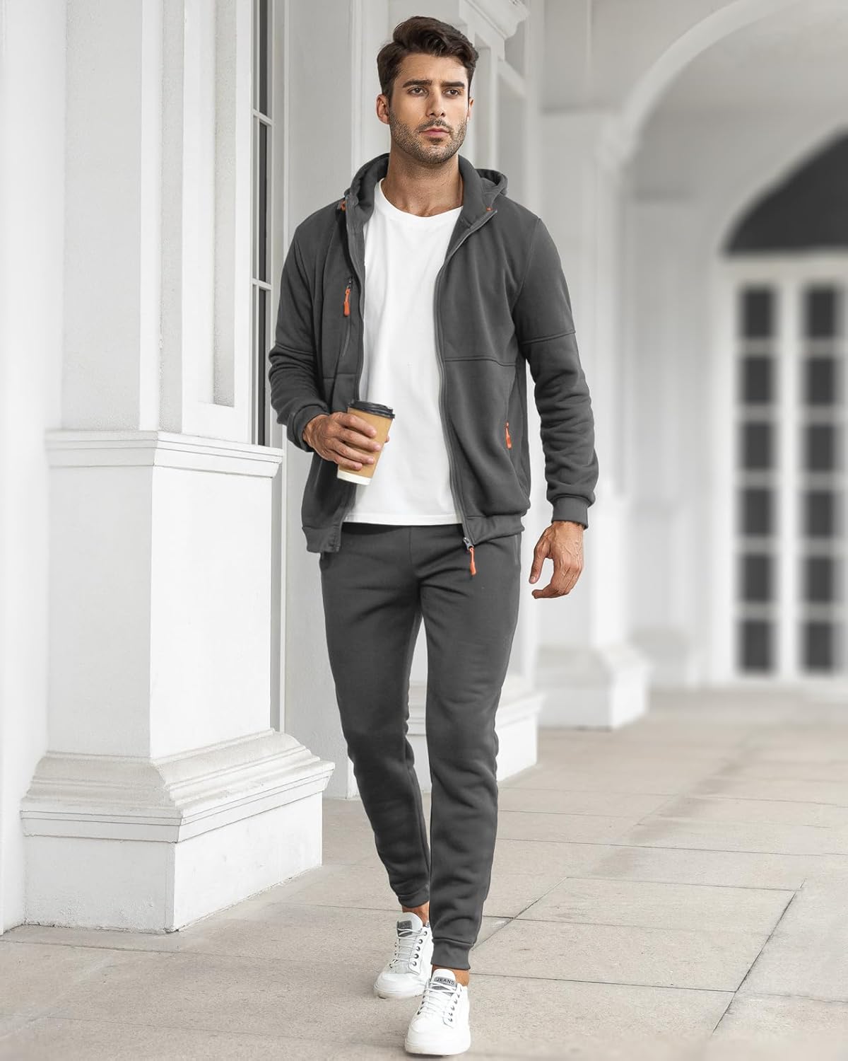 Man in gray hoodie and pants holding a coffee cup in an architectural setting