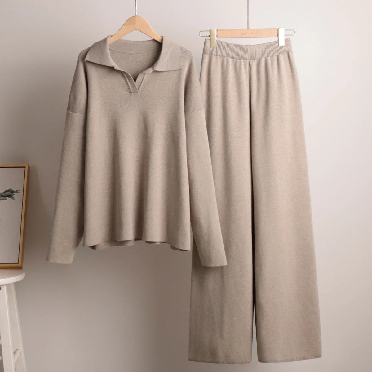 Beige long-sleeve top and pants set hanging on wooden hangers against a plain wall.
